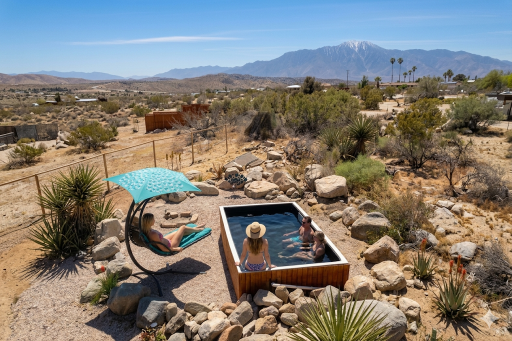 Desert yard with a wooden hot tub containing three people; a person sunbathing under a turquoise umbrella nearby.
